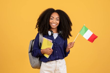 Téléchargez les photos : Joyeux jeune étudiant aux cheveux bouclés, tenant drapeau italien et cahiers, portant un casque autour de son cou, exsudant l'amour pour l'apprentissage et la culture sur fond jaune - en image libre de droit