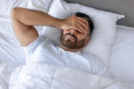 Photo for Sleepy and stressed man in bed covering face with hand, struggling with symptoms of hangover or fever at home bedroom. Guy suffering from chronic headaches and insomnia lying indoor - Royalty Free Image
