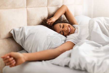 Téléchargez les photos : Réveil matinal. Happy Rested Jeune dame noire se réveillant après bien dormir et étirer les mains, allongé dans un lit confortable dans l'intérieur de la chambre à coucher confortable, souriant avec les yeux fermés - en image libre de droit