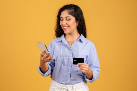 Photo for Smiling latin millennial brunette woman in casual typing on smartphone, use credit card, enjoy online shopping, isolated on orange studio background. Sale, finance, banking app - Royalty Free Image