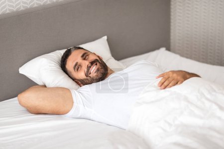 Téléchargez les photos : Homme européen détendu se prélassant joyeusement dans un lit confortable, se réveillant tranquillement dans une chambre moderne confortable, souriant alors qu'il était allongé confortablement sous une couverture blanche, profitant d'une bonne matinée - en image libre de droit