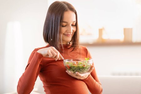 Téléchargez les photos : Bonne femme enceinte assise sur le canapé et dégustant une salade fraîche pour le déjeuner à la maison. Joyeux attente future maman ayant bol de légumes en bonne santé dans le salon intérieur. Nutrition et grossesse - en image libre de droit