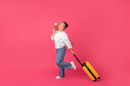 Foto de Concepto de viaje. Retrato de mujer asiática joven feliz con maleta y pasaporte caminando sobre fondo de estudio rosa, alegre mujer coreana lista para las vacaciones, longitud completa, espacio de copia - Imagen libre de derechos