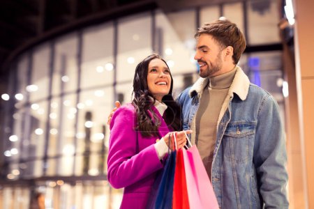 Photo for Young couple holds shopper bags full of Valentines Day gifts and embracing near modern city mall outside. Spouses celebrating their love purchasing gifts on winter evening. Free space - Royalty Free Image