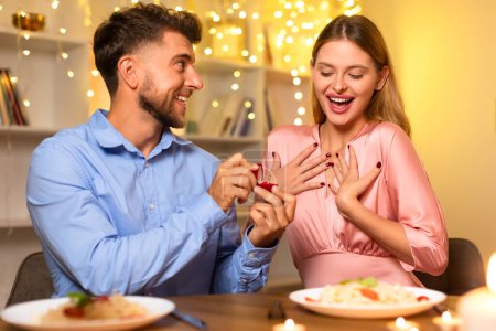 Photo for Excited young woman in pink blouse expressing surprise as man in blue shirt presents engagement ring at candlelit dinner with pasta and wine - Royalty Free Image