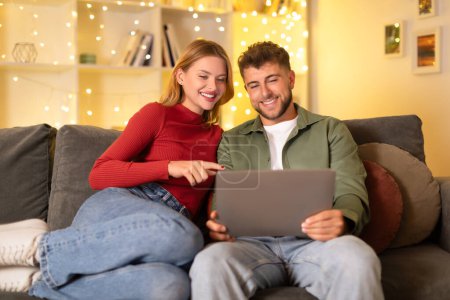 Photo for Cheerful young european couple browsing on digital tablet, lounging on comfortable couch in room illuminated by warm fairy lights - Royalty Free Image
