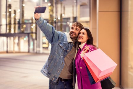 Photo for Millennial couple joyfully taking a selfie on smartphone outside city mall, posing with shopping bags, capturing their festive shopping spree on winter evening using cell phone app. - Royalty Free Image