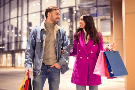 Photo for Millennial couple enjoying winter shopping trip, carries bags full of Christmas gifts, sharing joyful moment walking outside a mall together in evening. New Year And Valentines Day sales offer - Royalty Free Image