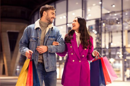 Photo for Contented couple shopping during winter spree and sales, carrying bags of New Years purchases, walking near a mall with festive lights outdoors, spending evening in city. - Royalty Free Image