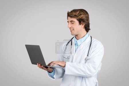 Téléchargez les photos : Médecin masculin confiant en manteau blanc avec stéthoscope tapant sur ordinateur portable isolé sur fond gris, studio. Consultation en ligne, service médical à distance, soins de santé - en image libre de droit