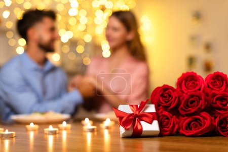 Photo for Focused on gift and bouquet of red roses, with joyful couple blurred in the background, celebrating romantic candlelit dinner at home, closeup - Royalty Free Image