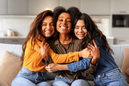 Foto de Tres novias diversas abrazándose sentadas en el sofá en el interior de casa. Grupo de jóvenes sonrientes celebrando su amistad pasando tiempo juntos, abrazando posando en interiores - Imagen libre de derechos