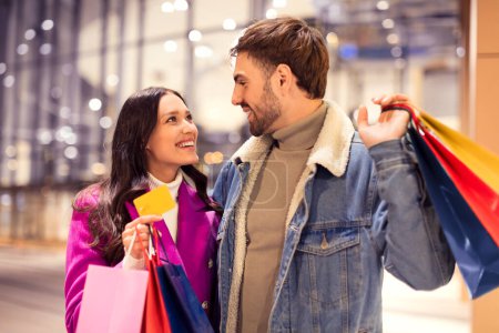 Photo for Lady shows credit card to man happy to start winter holiday shopping, hugging outside city mall at night. Couple in winter jackets finding the perfect gifts for Christmas and Valentines Day - Royalty Free Image