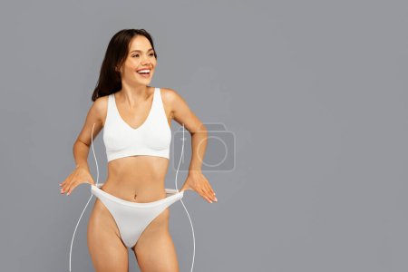 Photo for Happy asian millennial woman in white underwear, laughing and playful stretching underpants, looking at free space with abstract fat figure. Playfulness and positive body, weight loss result - Royalty Free Image