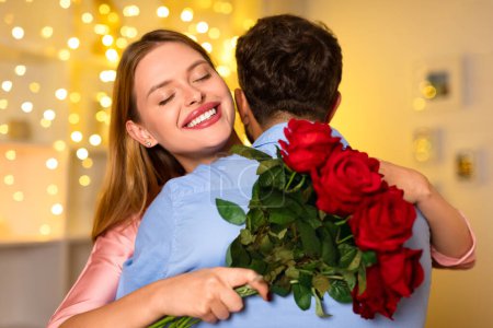 Téléchargez les photos : Femme avec un sourire lumineux étreignant l'homme par derrière, serrant bouquet de roses rouges, avec des lumières de ficelle scintillantes fournissant un fond confortable - en image libre de droit