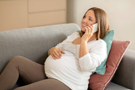 Photo for Beautiful pregnant woman talking on cellphone and embracing her belly while sitting on couch at home, happy expectant mother enjoying pleasant conversation and relaxing in living room, free space - Royalty Free Image