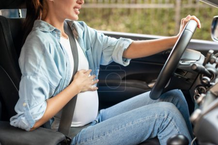 Photo for Motherhood, responsible driving. Cropped shot of pregnant lady driver navigating car vehicle and touching her growing belly, feeling discomfort during ride. Side view, closeup - Royalty Free Image