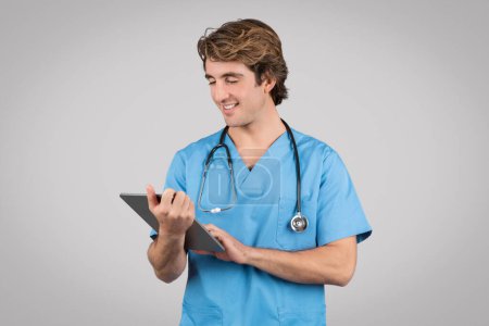 Foto de Feliz médico varón de capa azul trabajando en tableta digital aislada sobre fondo gris. Profesional médico, consulta de terapeuta a distancia con gadget - Imagen libre de derechos
