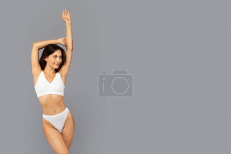 Photo for A fit young woman in a white sports bra and panties stretches her arms above her head, looking away with a subtle smile against a plain grey backdrop, suggesting health and simplicity - Royalty Free Image