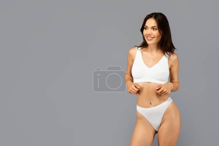 Photo for Cheerful european young woman in white sports bra and underwear, smiling and looking to empty space, enjoy beauty care, isolated on grey backdrop. Active lifestyle and sport, body care - Royalty Free Image