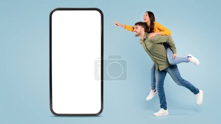 Téléchargez les photos : Joyeux couple dans la pose de piggyback pointant, à côté de la maquette géante du smartphone, sur fond bleu, plein de vie et de mouvement, espace libre, bannière - en image libre de droit