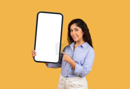 Photo for Smiling latin millennial brunette woman point finger at big smartphone with empty screen, isolated on orange studio background. Ad and offer, work, study app, social networks recommendation - Royalty Free Image