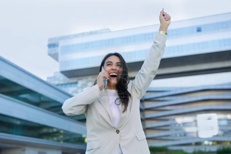 Photo for Business Success. Joyful Latin Businesswoman Talking On Phone Shaking Raised Arms, Celebrating Achievement And Great Corporate News, Standing Outdoors Modern Office Center - Royalty Free Image