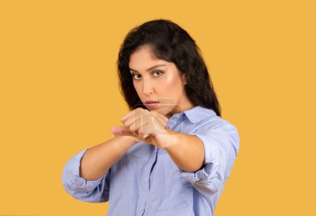 Photo for Resolute young woman with long curly hair, wearing a light blue shirt, stands with her fists up in a defensive stance, ready to fight or defend, against a yellow background - Royalty Free Image
