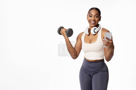 Photo for Young athletic African American woman with phone and headphones holding dumbbell, stands over white studio background. Fitness and strength, online training application. Copy space - Royalty Free Image