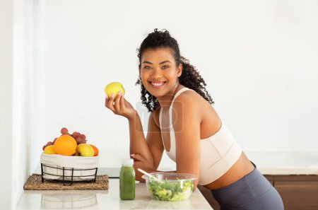 Foto de Dieta adelgazante saludable. Joven y alegre mujer de fitness sosteniendo una fruta fresca de manzana, sonriendo a la cámara posando en su cocina en casa. Estilo de vida del concepto de salud y nutrición - Imagen libre de derechos
