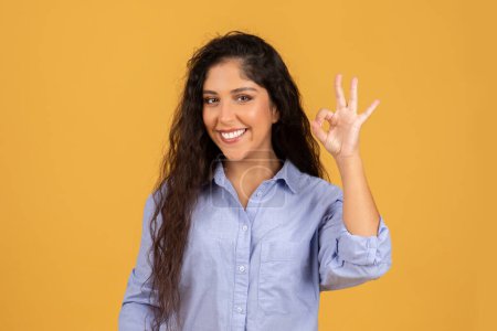 Photo for Smiling latin millennial brunette lady in casual show ok sign with hand, recommend sale, isolated on orange studio background. Ad and offer, attention good news, approve gesture - Royalty Free Image