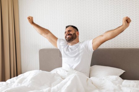 Téléchargez les photos : Européen barbu gars se réveillant dans la chambre moderne et étire les bras avec un sourire heureux, assis dans le lit avec des draps blancs. Une routine matinale confortable et détendue à la maison concept - en image libre de droit