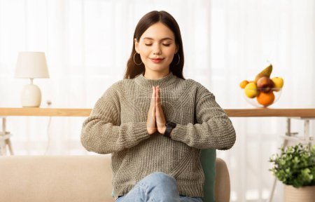 Photo for Calm young brunette woman sitting on couch with closed eyes and smile on her face, concentrating or meditating, praying in the morning, holding hands in namaste gesture, home interior - Royalty Free Image