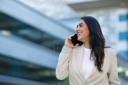 Téléchargez les photos : Femme d'affaires hispanique avoir une conversation sur smartphone, parler avec le client à l'extérieur du centre-ville. Heureuse femme entrepreneure millénaire qui gère le travail et la communication en déplacement. Espace de copie - en image libre de droit