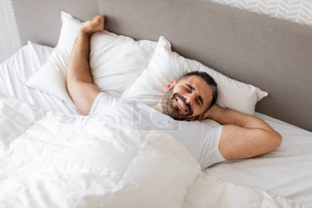 Photo for Smiling middle aged caucasian man stretching his arms, wakes up on cozy bed with white linens. Guy enjoying lazy morning awakening in modern bedroom at home. Healthy restful sleep - Royalty Free Image