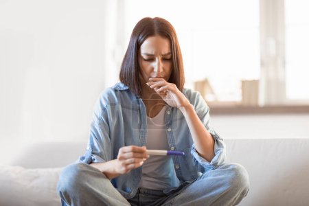 Photo for Upset disappointed young european lady holds negative pregnancy test, sits on sofa alone in living room indoor. Infertility, female gynecology health issues, unwanted pregnancies and loneliness - Royalty Free Image