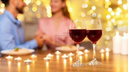 Foto de Concéntrate en dos copas de vino tinto con una pareja borrosa cogida de la mano y un ambiente romántico a la luz de las velas en el fondo, creando un ambiente gastronómico íntimo. - Imagen libre de derechos