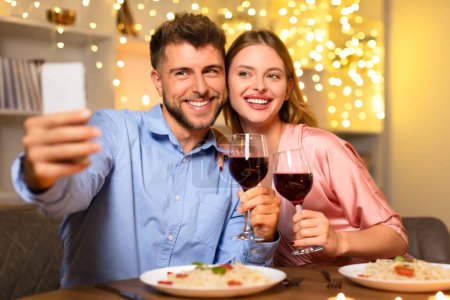 Photo for Joyful young couple sharing a moment, taking a selfie with glasses of red wine during a romantic candlelit dinner, celebrating together - Royalty Free Image