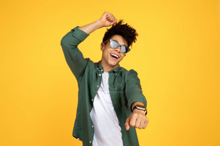 Photo for Carefree stylish young black guy wearing sunglasses with smart watch on his wrist dancing and singing isolated on yellow studio background, have fun, enjoying holiday or vacation - Royalty Free Image