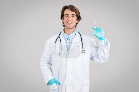 Photo for Positive man doctor in white coat, protective gloves using syringe, recommending vaccination, standing isolated on gray background. Medicine and injection, health care - Royalty Free Image