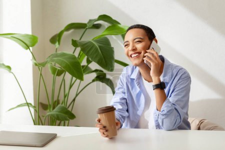 Photo for Smiling young lady with short hair, with smartwatch holding coffee cup in hand, talking on mobile phone, with laptop and indoor plants in background. Remote work and communication - Royalty Free Image