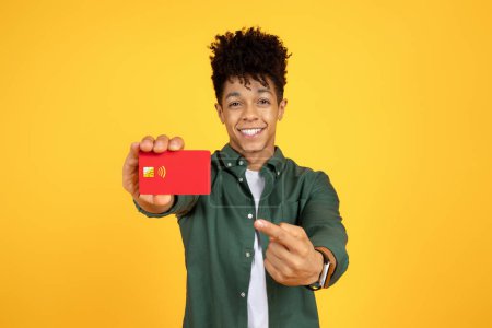 Foto de Alegre guapo joven afroamericano hombre apuntando a la tarjeta de crédito del banco rojo y sonriente, cliente satisfecho demostrando producto bancario sobre fondo de estudio amarillo. Financiación, préstamo, concepto de ahorro - Imagen libre de derechos