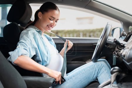 Téléchargez les photos : Jeune femme enceinte dans sa voiture réglage de la ceinture de sécurité pour le confort et la sécurité, posant dans le siège du conducteur, se préparant pour la promenade à travers la ville. Voyage du concept de maternité. Vue latérale - en image libre de droit