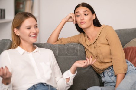 Envy In Friendship. Jealous Lady Listening To Her Cheerful Friend Talking And Bragging About Her Great Life Sitting On Couch At Home, Rolling Eyes During Conversation. Selective Focus