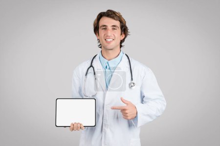 Foto de Alegre joven doctor apuntando con el dedo a la tableta con pantalla blanca en blanco sobre fondo gris, maqueta. Gran anuncio médico y oferta, tratamiento con terapeuta remoto - Imagen libre de derechos