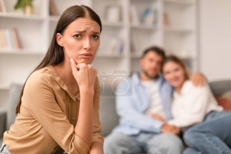 Photo for Jealousy. Sad Young Woman Looking At Camera While Her Ex Boyfriend Hugging New Girlfriend Sitting On Couch Indoors. Love Triangle And Envy In Friendship With Married Friends. Selective Focus - Royalty Free Image