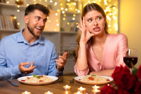 Téléchargez les photos : Femme tenant la tête dans l'ennui tandis que l'homme souriant parle lors d'un dîner romantique aux chandelles avec des pâtes et du vin, des roses floues au premier plan - en image libre de droit