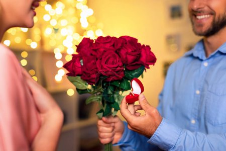 Foto de En una habitación cálidamente iluminada, el hombre sorprende a su pareja con una propuesta de matrimonio, presentando un brillante anillo de compromiso junto a un ramo de rosas rojas. - Imagen libre de derechos
