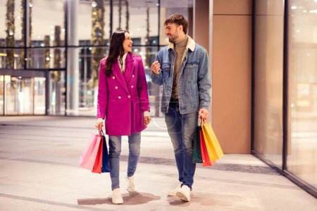 Photo for Winter shopping offer. Joyful European husband and wife carry shopper bags filled with New Years goodies, strolling past a city mall under evening city lights. Full length shot - Royalty Free Image