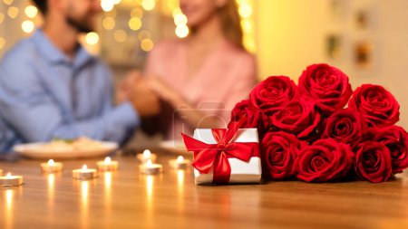 Photo for White gift box tied with red ribbon lies next to stunning bouquet of red roses on table, where couple is deep in conversation over romantic candlelit dinner - Royalty Free Image